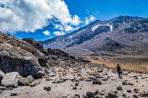 A person walks on a rocky trail with sparse vegetation, set against a backdrop of a snow-capped mountain under a bright blue sky with scattered clouds.